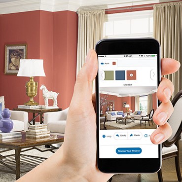 Light brick-colored room with white couches and decorations with brown tables and purple accents with person holding phone taking picture of room to decide colors